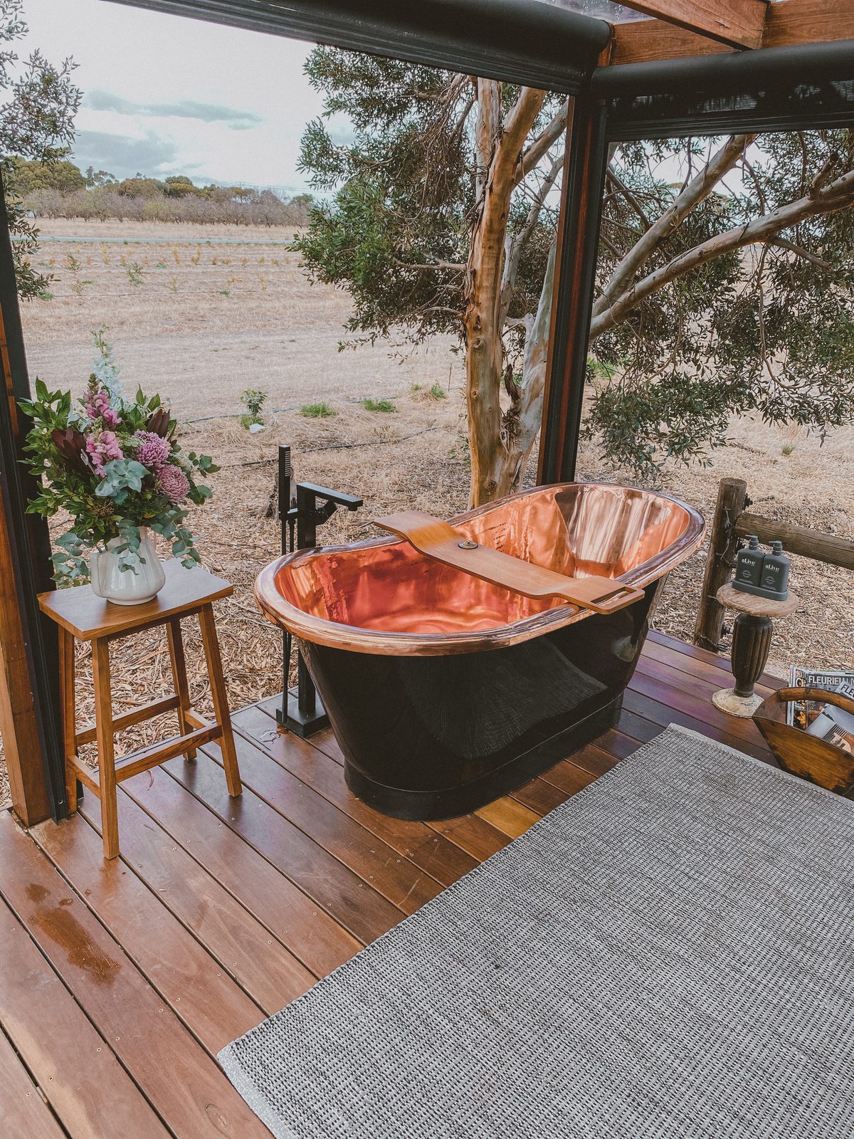 The copper bath on the deck under the pergola, flowers on a stool beside it and the vineyard beyond